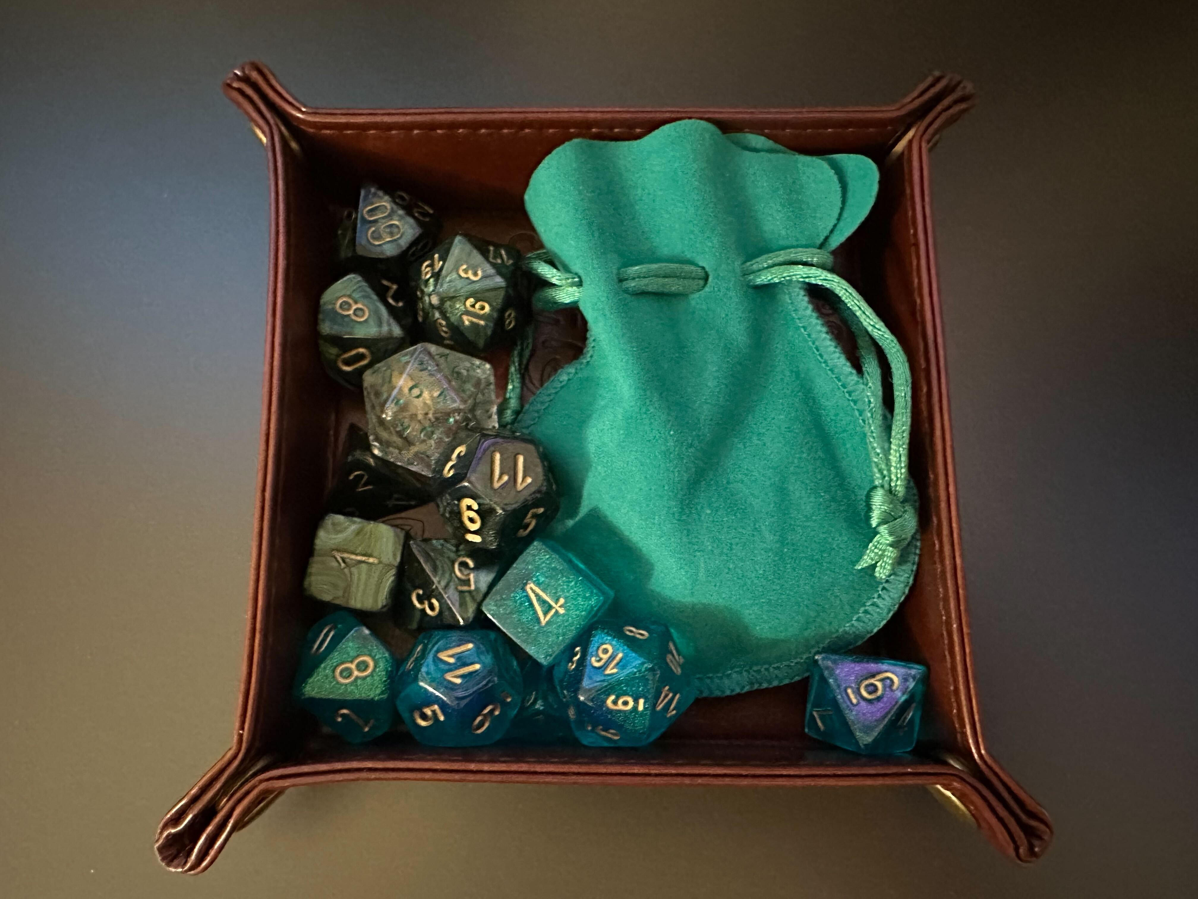 Photo of D4s, D6s, D8s, D20s in various shades of green in a fancy brown box, next to a soft looking emerald bag.